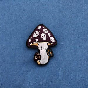 May include: A purple and white mushroom patch with a skull design. The mushroom is embroidered on a dark blue fabric.
