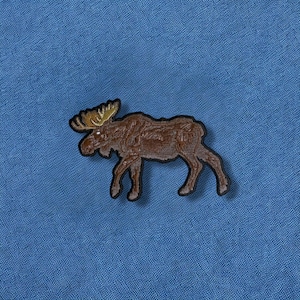 May include: A brown moose patch with a detailed design, featuring antlers and a textured body. The patch is set against a blue fabric background, creating a contrast that highlights the moose's form. This patch is suitable for various applications.