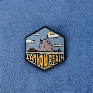 May include: A hexagonal embroidered patch with a blue sky, white clouds, a yellow sun, a gray mountain, and green trees. The text "Cannon Beach" is embroidered in yellow on a black background.