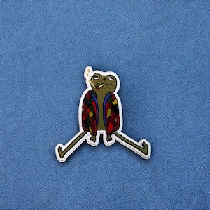 May include: A green frog wearing a red, blue, and yellow patterned jacket with white outlines. The frog is sitting with its legs spread out and has a cigarette in its mouth.