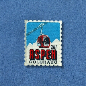 May include: Embroidered patch featuring a red ski gondola against a blue sky and white mountain backdrop. The patch reads "Aspen Colorado" in red and black lettering, with "Ski!" above the gondola.