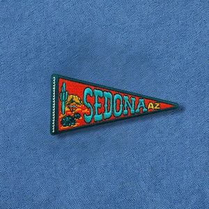 May include: A red, teal, and gold Sedona AZ pennant patch. The patch features a teal cactus and mountain design with the word "SEDONA" in teal lettering. The patch is bordered in teal and has a sawtooth edge.