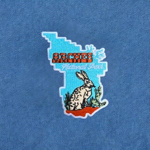 May include: A blue fabric surface featuring an embroidered patch shaped like a jagged map. The patch displays the orange text ARCHES, the white text National Park, and a detailed illustration of a rabbit sitting near a green cactus.