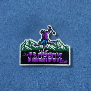 May include: Embroidered patch featuring a skier in purple and blue, set against a backdrop of green mountains. The patch reads "Ski Jackson Hole" in purple letters on a black background, with a white border. The patch is on a blue fabric.