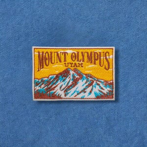 May include: Embroidered patch featuring Mount Olympus in Utah. The patch has a rectangular shape with a yellow background and a mountain range design in shades of brown, white, and blue. The text "MOUNT OLYMPUS UTAH" is arched across the top.