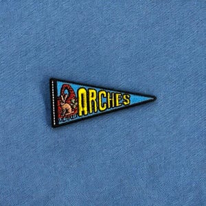 May include: A blue and red pennant with the word "ARCHES" in yellow embroidered letters. The pennant has a small image of a rabbit and the word "MOAB" in white.
