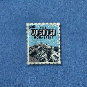 May include: Embroidered patch featuring a mountain landscape and the text "Visit The Wasatch Mountains." The patch has a light blue background with a white border and is set against a blue fabric.