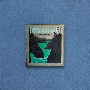 May include: Square patch with a silver border featuring an image of the Oregon Coast. The image depicts a turquoise body of water surrounded by dark cliffs and trees. The text "OREGON COAST Natural Bridges" is printed at the top.