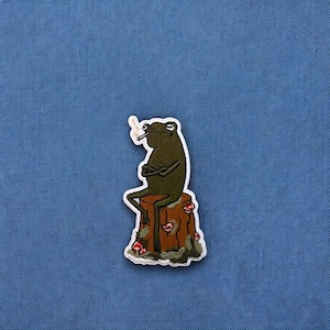 May include: A green frog with a cigarette in its mouth sits on a brown tree stump with red mushrooms. The frog is wearing a white outline and is sitting with its arms crossed.