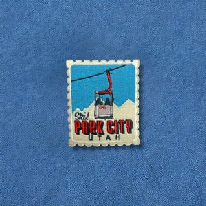 May include: Embroidered patch featuring a ski lift design with the words "Ski! Park City Utah" in red and black. The patch is in a postage stamp shape with a cream-colored border, set against a blue fabric background.
