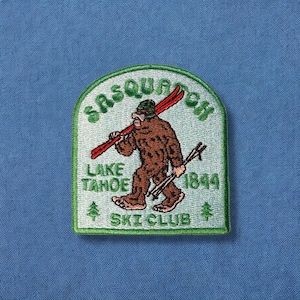 May include: Green embroidered patch with a cartoon Sasquatch holding skis. The text reads "Sasquatch Lake Tahoe Ski Club 1844".