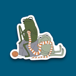 May include: Two green frogs, one knitting an orange and white striped scarf for the other frog.