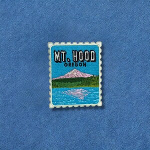 May include: Embroidered patch featuring a scenic view of Mt. Hood in Oregon. The design includes a mountain, lake, and the text "MT. HOOD OREGON" in a postage stamp style. The patch has a white border and is set against a blue background.
