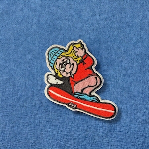 May include: A colorful embroidered patch of a cartoon character riding a surfboard. The character has blonde hair, a blue hat, and is wearing a red shirt. The surfboard is red and white.