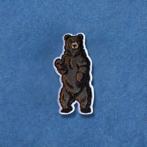 May include: Embroidered patch featuring a brown bear standing upright with a white border. The bear has detailed fur and is set against a blue background. This patch is suitable for jackets, bags, or other fabric items.