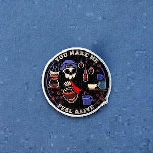 May include: A round, white embroidered patch with a black background. The patch features a skull wearing a purple beanie, pouring coffee into a cup. The text "You Make Me Feel Alive" is written around the edge of the patch.