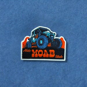 May include: Embroidered patch featuring a blue off-road vehicle with large tires. The patch has the words "Ride Moab Utah" in orange and white. The patch is on a blue background.