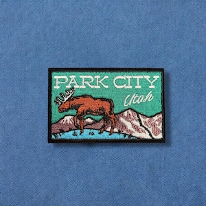 May include: Embroidered patch featuring a moose in front of mountains, with the text "PARK CITY Utah" in white. The patch has a black border and is set against a teal background. The patch is on a blue fabric.