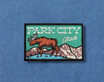 May include: Embroidered patch featuring a moose in front of mountains, with the text "PARK CITY Utah" in white. The patch has a black border and is set against a teal background. The patch is on a blue fabric.