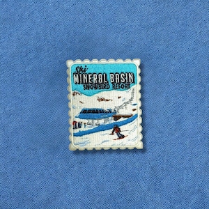 May include: A rectangular embroidered patch with a scalloped edge featuring a snowy mountain scene. The text reads Ski MINERAL BASIN SNOWBIRD RESORT. The design includes a blue ski lift, a skier in red, and white snow against a light blue sky.