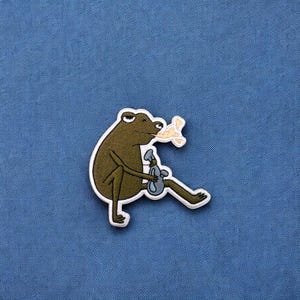 May include: Embroidered patch featuring a cartoon frog in a relaxed pose, holding a spray bottle and a yellow object. The frog is olive green with a white border. The patch is on a blue background.