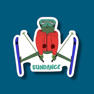 May include: A sticker featuring a cartoon frog wearing ski goggles and a red jacket, holding ski poles, and skis. The word "SUNDANCE" is written in green below the frog.