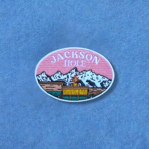 May include: An oval embroidered patch featuring a pink sky above white snow-capped mountains. The text JACKSON HOLE is stitched in white above a brown building with the text COWBOY BAR in yellow. The patch is set against a light blue fabric.