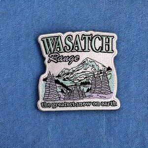 May include: Embroidered patch featuring the words "WASATCH Range" above a mountain scene with pine trees. The text "the greatest snow on earth" is at the bottom. The patch has a white border and is on a blue background.