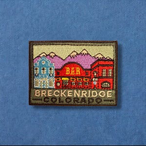 May include: Embroidered patch with a green border, depicting a stylized image of a town with a building that says "BAR" and the text "BRECKENRIDGE COLORADO" below.
