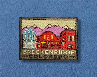 May include: Embroidered patch with a green border, depicting a stylized image of a town with a building that says "BAR" and the text "BRECKENRIDGE COLORADO" below.