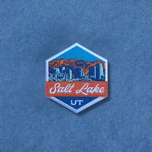 May include: Embroidered patch with a hexagonal shape featuring a Salt Lake City design. The patch has a blue and orange color scheme, with the words "Salt Lake UT" in white script. The patch is on a blue fabric background.