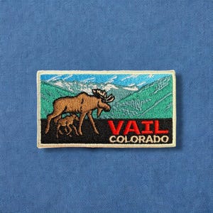 May include: A white patch with a brown and white moose and a calf in a green mountain landscape. The text "VAIL COLORADO" is in red letters.