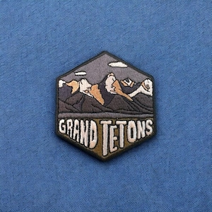 May include: Embroidered patch featuring a mountain range design with the text "GRAND TETON". The patch is in a hexagonal shape with a black border, and the mountains are depicted in shades of gray, brown, and white. The text is in a stylized font.