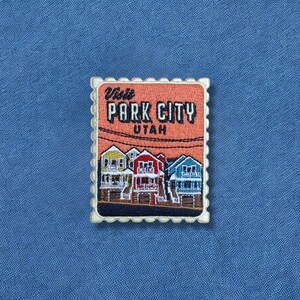 May include: A rectangular embroidered patch shaped like a postage stamp with a scalloped edge. It features a peach-colored sky with the text "Visit PARK CITY UTAH" above a row of colorful houses in yellow, red, and blue against a dark base.