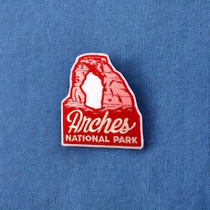 May include: Red and white embroidered patch with a depiction of Delicate Arch in Arches National Park, Utah. The text "Arches National Park" is below the arch.