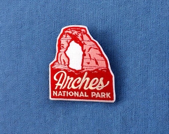 May include: A red and white embroidered patch featuring the iconic arches of Arches National Park. The patch has a white border and the words "Arches NATIONAL PARK" in a cursive font.