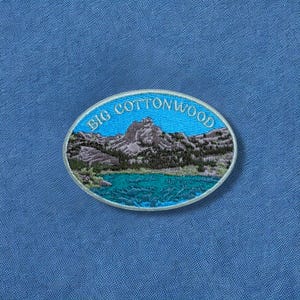 May include: An embroidered patch with an oval shape, featuring a mountain landscape and a lake. The patch has the words "BIG COTTONWOOD" in a curved font at the top. The background is a solid blue color.