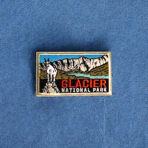 May include: A brown and beige embroidered patch with a white mountain goat standing on a rock. The patch has the text "Glacier National Park" in red and blue lettering.