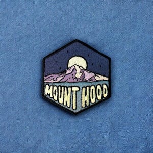 May include: A hexagonal patch featuring a mountain landscape under a night sky with a full moon. The patch reads "MOUNT HOOD" in a stylized font, with the mountain in shades of purple and white.