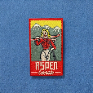 May include: Red, white, and yellow embroidered patch with a woman skiing down a mountain. The patch says "Aspen - Colorado" in a white font.