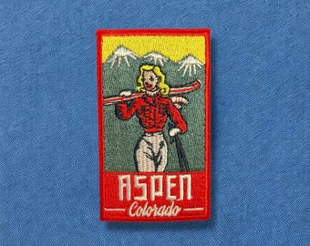 May include: Embroidered patch featuring a skier in a red jacket and white pants, carrying skis. The patch has a red border and includes the text "ASPEN - Colorado" with a mountain backdrop and yellow sky.
