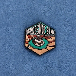 May include: A black embroidered patch with a hexagonal shape. The patch features a colorful illustration of Horseshoe Bend, a famous geological formation in Arizona, with the text "Horseshoe Bend" above the illustration.
