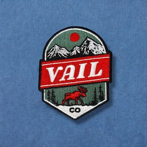 May include: A green, red, and white embroidered patch with a mountain scene and a moose. The patch says "Vail CO" in white text.