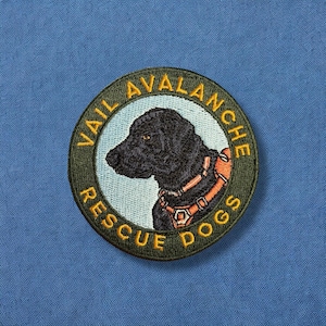 May include: A green and yellow embroidered patch with the text "Vail Avalanche Rescue Dogs" and a black Labrador Retriever dog with a red and orange harness.