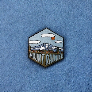 May include: A hexagonal embroidered patch with a mountain scene and the text "MOUNT RAINIER" in green and white lettering. The scene depicts a mountain with snow on the peak, a blue sky with white clouds, and a small orange sun.