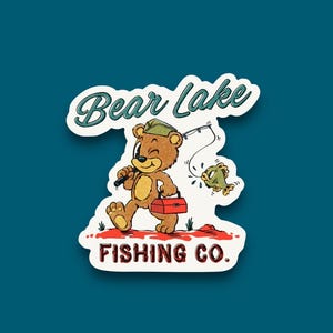 May include: A cartoon bear wearing a green hat and carrying a red tackle box walks with a fishing rod and a green fish. The text "Bear Lake Fishing Co." is below the bear.