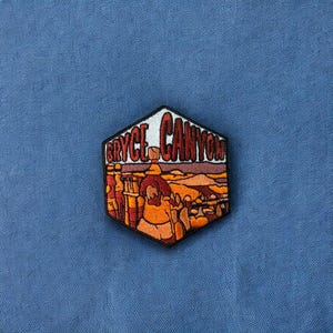May include: A hexagonal embroidered patch with the text "Bryce Canyon" in red and white lettering. The patch features a colorful illustration of a desert landscape with people standing on a rock formation.
