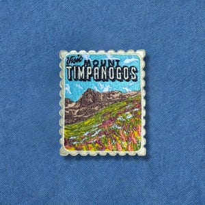 May include: Embroidered patch featuring a mountain landscape with the text "Visit Mount Timpanogos." The patch has a postage stamp design with a blue sky, brown mountain, and green and purple vegetation.