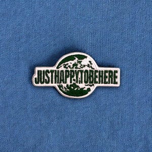 May include: A white and green embroidered patch with the text "Just Happy to Be Here" and a green globe design.