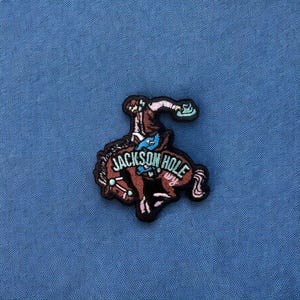 May include: Embroidered patch featuring a brown horse with a cowboy riding it. The horse is bucking and the cowboy is holding onto the reins. The patch has the text "JACKSON HOLE WY" in a white circle around the horse and rider.